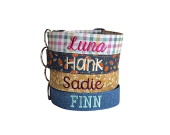 May include: A stack of four fabric pet collars with metal D-rings. The top collar is a plaid pattern with the name Luna embroidered in pink. The second is navy with pumpkins and footballs, embroidered with Hank. The third is mustard with floral patterns, embroidered with Sadie. The bottom is blue herringbone, embroidered with FINN.