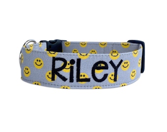 May include: A light gray pet collar featuring a repeating pattern of yellow smiley faces. The name Riley is embroidered in bold black lettering across the center. The collar includes a black plastic side-release buckle and a silver metal D-ring.