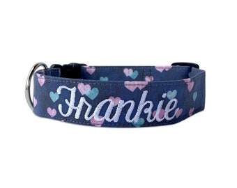 May include: A dark blue pet collar featuring a repeating pattern of small light pink and teal hearts. The name Frankie is embroidered in white cursive script across the center. The collar includes a silver metal D-ring and a black plastic buckle.