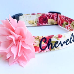 May include: A pet collar featuring a floral pattern in shades of red, pink, and green, accented with a large, layered light pink fabric flower. The collar includes a black plastic buckle and is embroidered with the name Chevelle in dark blue.