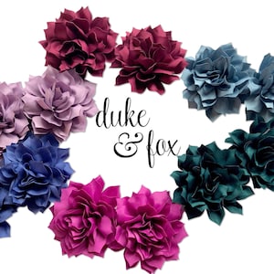 May include: A collection of layered fabric flowers in various colors including burgundy, lavender, navy blue, magenta, and dark teal, arranged in a circular pattern around the black text "duke & fox" centered on a plain white background.