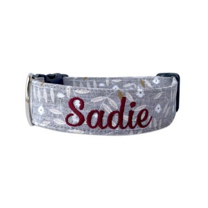 Op de afbeelding: Grijze hondenriem met een bloemenpatroon met witte madeliefjes en gouden accenten. De naam "Sadie" is geborduurd in bordeauxrode draad. De halsband heeft een zwarte plastic gesp en een zilveren metalen lus.