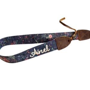 May include: A navy blue floral patterned guitar strap with the name "Anel" embroidered in white. The strap has brown leather accents and a leather strap for attachment.