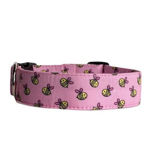 May include: Pink dog collar featuring a repeating pattern of yellow and black cartoon bees. The collar has a black plastic buckle and a silver metal loop for attaching a lead. The collar is made of fabric.