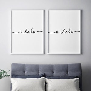 May include: Two framed wall art prints with the words "inhale" and "exhale" in black script. The prints are displayed above a bed with a blue headboard, white pillows, and a dark blue blanket. A vase of greenery sits on a side table.