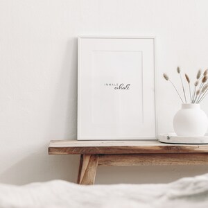 May include: White framed print with the text "inhale exhale" on a wooden shelf with a white vase and dried flowers.
