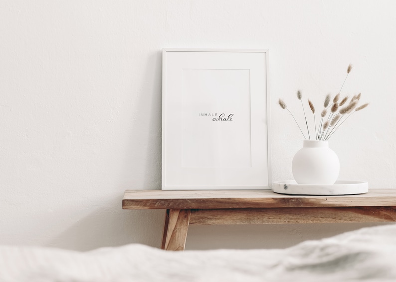 May include: White framed print with the text "inhale exhale" on a wooden shelf with a white vase and dried flowers.
