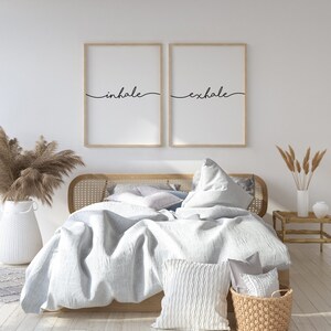May include: A bedroom scene featuring a bed with light blue bedding, decorative pillows, and a woven basket. Two framed prints above the bed read "inhale" and "exhale" in black script. A white vase with dried plants and a small wooden side table complete the decor.