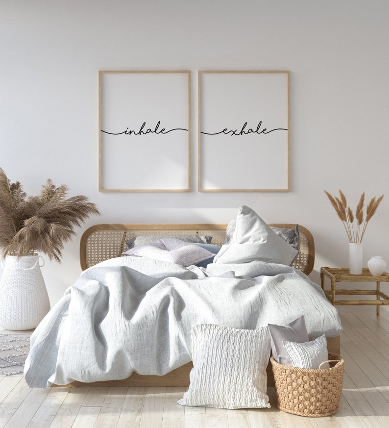 May include: A bedroom scene featuring a bed with light blue bedding, decorative pillows, and a woven basket. Two framed prints above the bed read "inhale" and "exhale" in black script. A white vase with dried plants and a small wooden side table complete the decor.