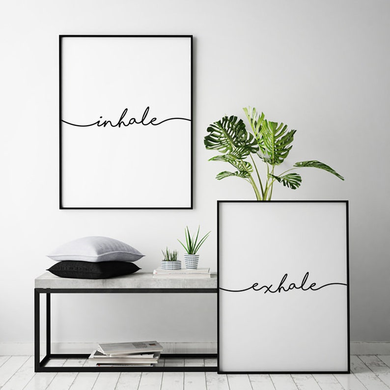 May include: Two framed minimalist art prints with black frames. One print says "inhale" in a cursive font, and the other says "exhale". A bench with pillows, plants, and books is in front of the prints.