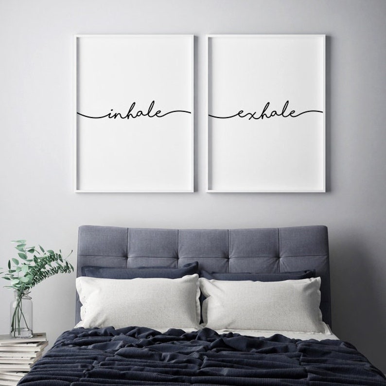 May include: Two framed wall art prints with the words "inhale" and "exhale" in black script. The prints are displayed above a bed with a blue headboard, white pillows, and a dark blue blanket. A vase of greenery sits on a side table.