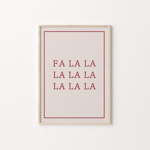 以下が含まれることがあります： ボルドー色の「FA LA LA LA LA LA LA」の文字がプリントされたフレーム入りプリント。テキストは、明るいベージュの背景に配置され、ボルドーのボーダーと明るい木製のフレームで囲まれています。お祝いのホリデーデコレーションです。