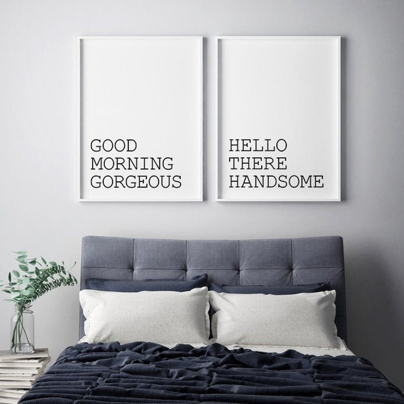 May include: Two white framed prints with black text on a white background. The left print says "Good Morning Gorgeous" and the right print says "Hello There Handsome". The prints are hanging above a gray headboard with a dark blue bedspread.