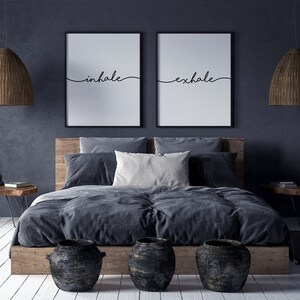 May include: A bedroom scene featuring a wooden bed frame with gray bedding and pillows. Two framed prints with the words "inhale" and "exhale" hang above the bed. Woven pendant lights and decorative vases complete the room's decor.