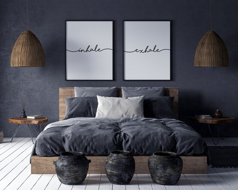 May include: A bedroom scene featuring a wooden bed frame with gray bedding and pillows. Two framed prints with the words "inhale" and "exhale" hang above the bed. Woven pendant lights and decorative vases complete the room's decor.