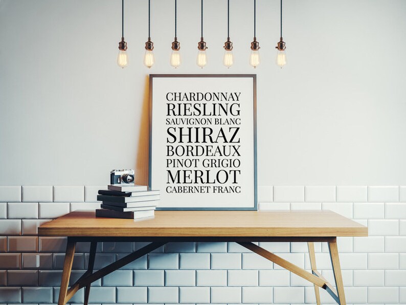 May include: A framed print with a list of wine varietals, including Chardonnay, Riesling, Sauvignon Blanc, Shiraz, Bordeaux, Pinot Grigio, Merlot, and Cabernet Franc. The print is hanging on a white tiled wall with a wooden table and a camera in front of it.