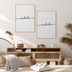 May include: Two framed wall art prints with the words "inhale" and "exhale" in black script. The prints are in gold frames and hang above a wooden cabinet. The room has a neutral color palette with natural textures.