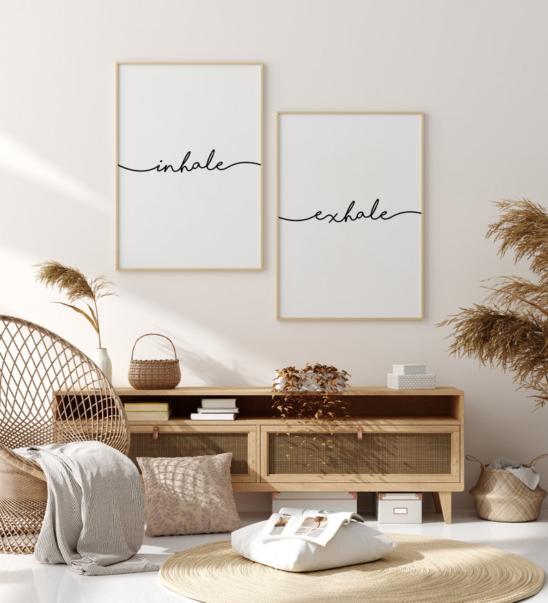 May include: Two framed wall art prints with the words "inhale" and "exhale" in black script. The prints are in gold frames and hang above a wooden cabinet. The room has a neutral color palette with natural textures.