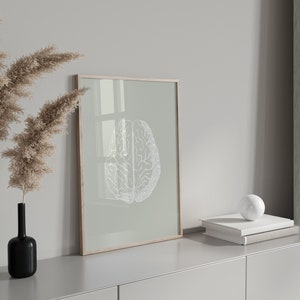 May include: A framed print of a white anatomical brain illustration on a light green background. The frame is wooden and has a light brown finish. The print is hanging on a white wall above a light gray dresser with a white ceramic sphere on top of a stack of books.