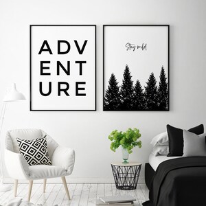 May include: Two black and white framed prints on a white wall. The print on the left says "ADVENTURE" in black text. The print on the right has a silhouette of a forest with the text "Stay wild" above it.