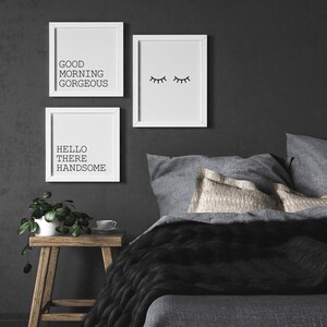 May include: Three white framed prints on a dark gray wall above a gray bed with black and gray bedding. The prints feature the text "Good Morning Gorgeous", "Hello There Handsome", and a simple line drawing of closed eyes.