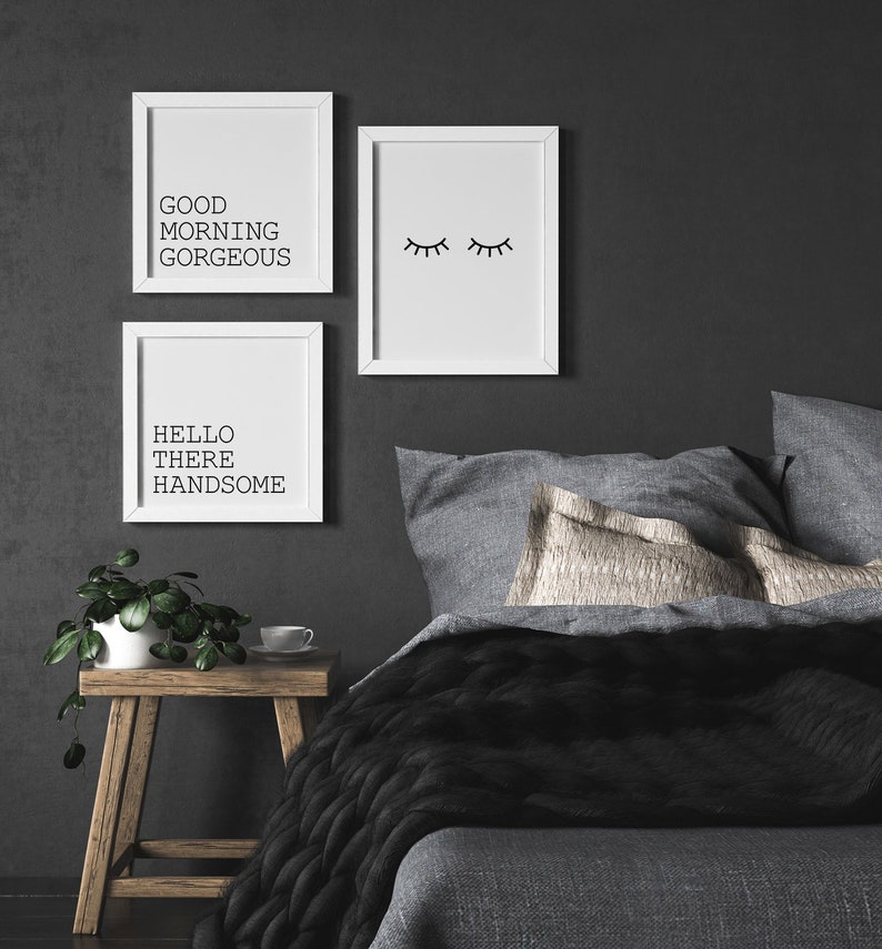 May include: Three white framed prints on a dark gray wall above a gray bed with black and gray bedding. The prints feature the text "Good Morning Gorgeous", "Hello There Handsome", and a simple line drawing of closed eyes.