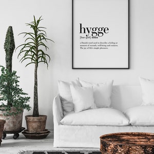 K&ouml;nnte beinhalten: Ein gerahmter Druck mit dem Wort "hygge" in schwarzer Schrift, definiert als d&auml;nisches Wort f&uuml;r W&auml;rme und Wohlbefinden. Der Druck h&auml;ngt &uuml;ber einem wei&szlig;en Sofa, mit Topfpflanzen und einem gemusterten Teppich.