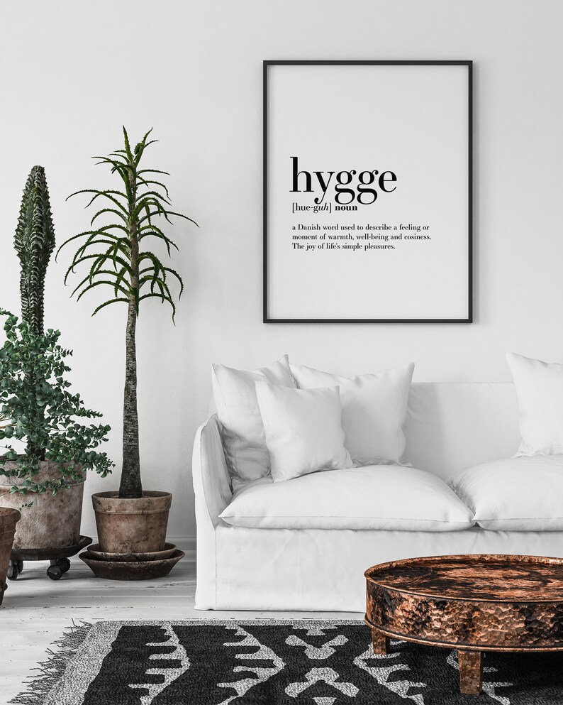 K&ouml;nnte beinhalten: Ein gerahmter Druck mit dem Wort "hygge" in schwarzer Schrift, definiert als d&auml;nisches Wort f&uuml;r W&auml;rme und Wohlbefinden. Der Druck h&auml;ngt &uuml;ber einem wei&szlig;en Sofa, mit Topfpflanzen und einem gemusterten Teppich.