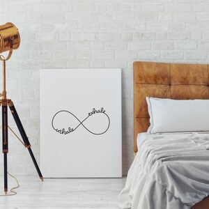 May include: A white canvas print featuring a black infinity symbol with the words "inhale" and "exhale" inscribed within it. The print is displayed against a white brick wall, above a brown leather headboard with white bedding.