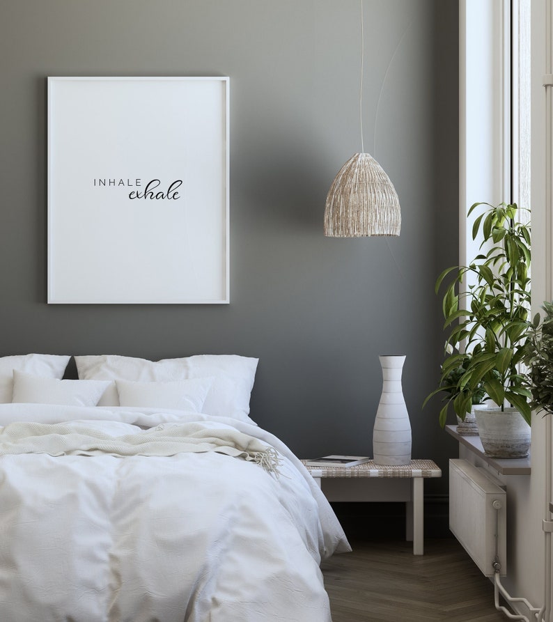 May include: A white framed print with the text "INHALE exhale" hanging on a gray wall above a white bed with white bedding and a white throw blanket. A white vase with a white and gray pattern is on a white wooden side table next to the bed. A woven pendant light hangs from the ceiling above the side table.
