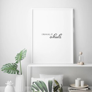 May include: White framed print with the text "INHALE exhale" in black cursive script. The print is on a white shelf with a white vase with a green leafy plant, a white pineapple, and a white candle holder. There are two green leaf patterned pillows on the shelf.