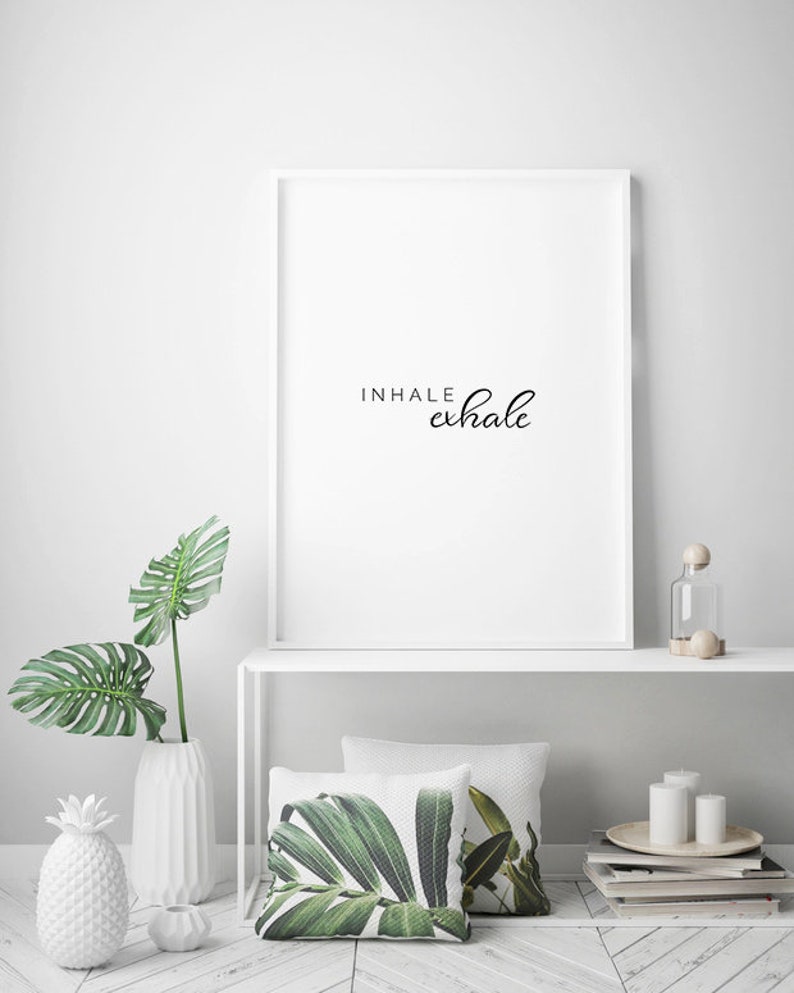 May include: White framed print with the text "INHALE exhale" in black cursive script. The print is on a white shelf with a white vase with a green leafy plant, a white pineapple, and a white candle holder. There are two green leaf patterned pillows on the shelf.