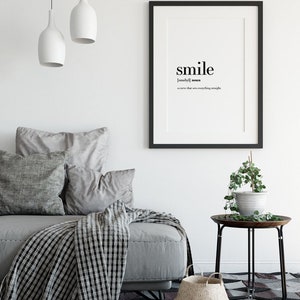 May include: Black and white typography print with the word "smile" defined as "a curve that sets everything straight". The print is framed in a black frame and hanging on a white wall above a gray couch with pillows and a black and white throw blanket.