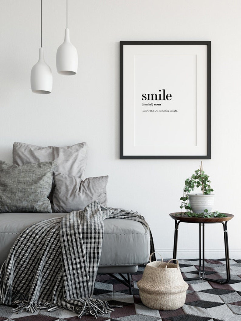May include: Black and white typography print with the word "smile" defined as "a curve that sets everything straight". The print is framed in a black frame and hanging on a white wall above a gray couch with pillows and a black and white throw blanket.