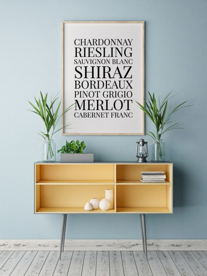 May include: A framed print with a white background and black text listing various types of wine: Chardonnay, Riesling, Sauvignon Blanc, Shiraz, Bordeaux, Pinot Grigio, Merlot, Cabernet Franc. The print is hanging on a light blue wall above a yellow shelf with a vase of greenery and a lantern.