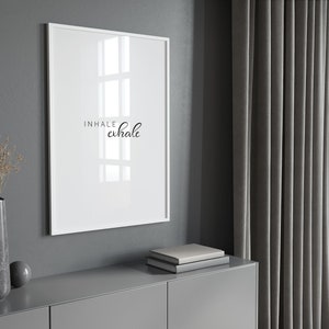 May include: A white framed print with the text "INHALE exhale" in black cursive lettering. The print is hanging on a gray wall above a gray dresser with a stack of books on top.