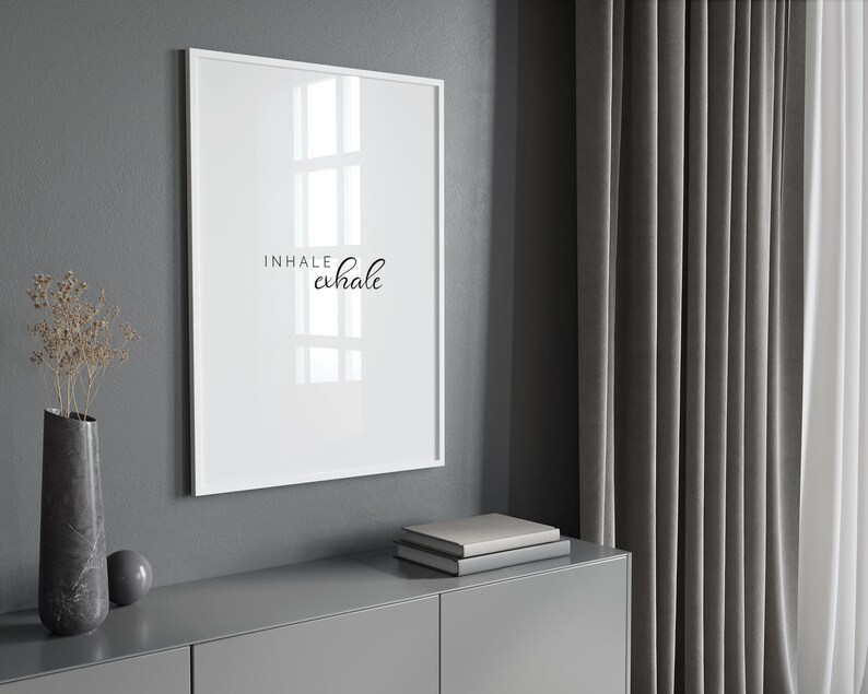 May include: A white framed print with the text "INHALE exhale" in black cursive lettering. The print is hanging on a gray wall above a gray dresser with a stack of books on top.