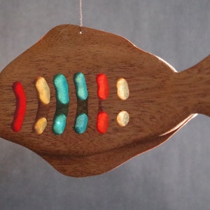Puede incluir: Un adorno de madera con forma de pez con detalles de vidrio de colores. El pez es marrón con una aleta caudal azul claro y una fila de piezas de vidrio verde, rojo, amarillo y azul a lo largo de su cuerpo.