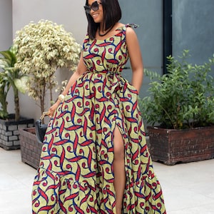 May include: A woman wearing a long, yellow, red, and blue patterned maxi dress with a side slit. The dress has a tie detail at the shoulders and a belt at the waist.