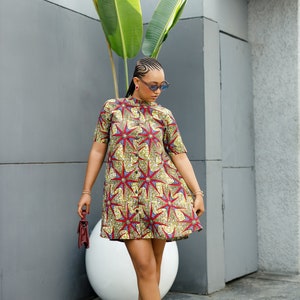 May include: A woman wearing a short-sleeved, yellow, red, and blue patterned dress with a button-up front. She is wearing gold sandals and sunglasses. She is holding a small, burgundy purse.