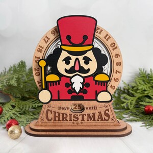 May include: Wooden Christmas countdown decoration featuring a red and black nutcracker soldier. The design includes a clock face with numbers and the text "Days 25 until Christmas". The base is made of layered wood.