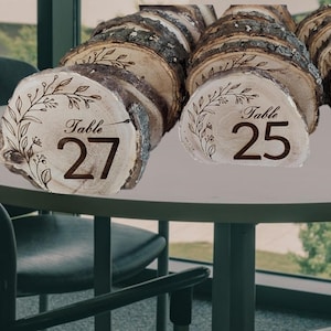 May include: Wooden table number holders with bark edges. Each holder displays a table number in a dark brown font, with decorative floral accents. The numbers visible are 25, 26, and 27. The holders are arranged on a round table.