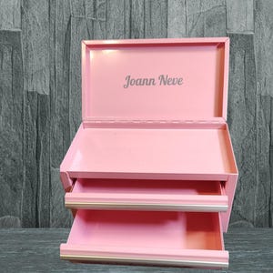 May include: A pink metal toolbox with two drawers and the name "Joann Neve" printed on the lid.