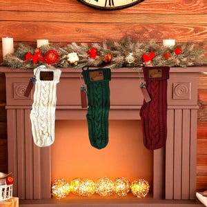 May include: Three cable-knit Christmas stockings in cream, green, and burgundy hang from a decorated mantel. Each stocking has a leather tag and a silver charm. The mantel is adorned with greenery, red bows, and candles, creating a festive holiday scene.