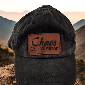 May include: Black baseball cap with a brown leather patch. The patch features the words "Chaos Coordinator" in a stylish font. The cap is made of a soft material and has a curved brim. The background shows a mountain landscape.