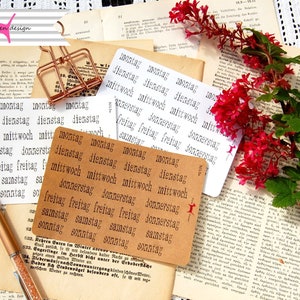 May include: A set of brown paper cards with the days of the week printed in German: Montag, Dienstag, Mittwoch, Donnerstag, Freitag, Samstag, Sonntag. The cards are arranged in a stack on a table with a pen and a book.