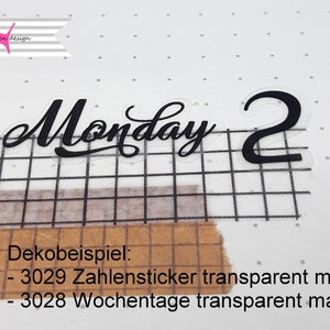 以下が含まれることがあります： 黒い文字で書かれた透明なステッカー。ステッカーには「Monday 2」と書いてあり、グリッドの背景に貼られています。ステッカーの下のテキストには「Dekobeispiel: - 3029 Zahlensticker transparent matt - 3028 Wochentage transparent matt」と書いてあります。