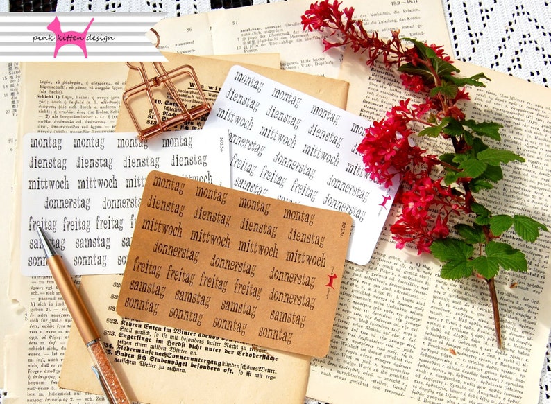 May include: A set of brown paper cards with the days of the week printed in German: Montag, Dienstag, Mittwoch, Donnerstag, Freitag, Samstag, Sonntag. The cards are arranged in a stack on a table with a pen and a book.