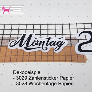 以下が含まれることがあります： 「Montag」という文字と数字「2」が書かれた白黒のステッカー。ステッカーは茶色と白のグリッドの背景の上にあります。「Dekobeispiel: - 3029 Zahlensticker Papier - 3028 Wochentage Papier」というテキストがステッカーの下にあります。