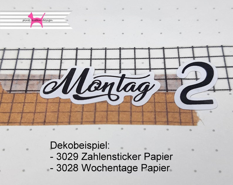 以下が含まれることがあります： 「Montag」という文字と数字「2」が書かれた白黒のステッカー。ステッカーは茶色と白のグリッドの背景の上にあります。「Dekobeispiel: - 3029 Zahlensticker Papier - 3028 Wochentage Papier」というテキストがステッカーの下にあります。
